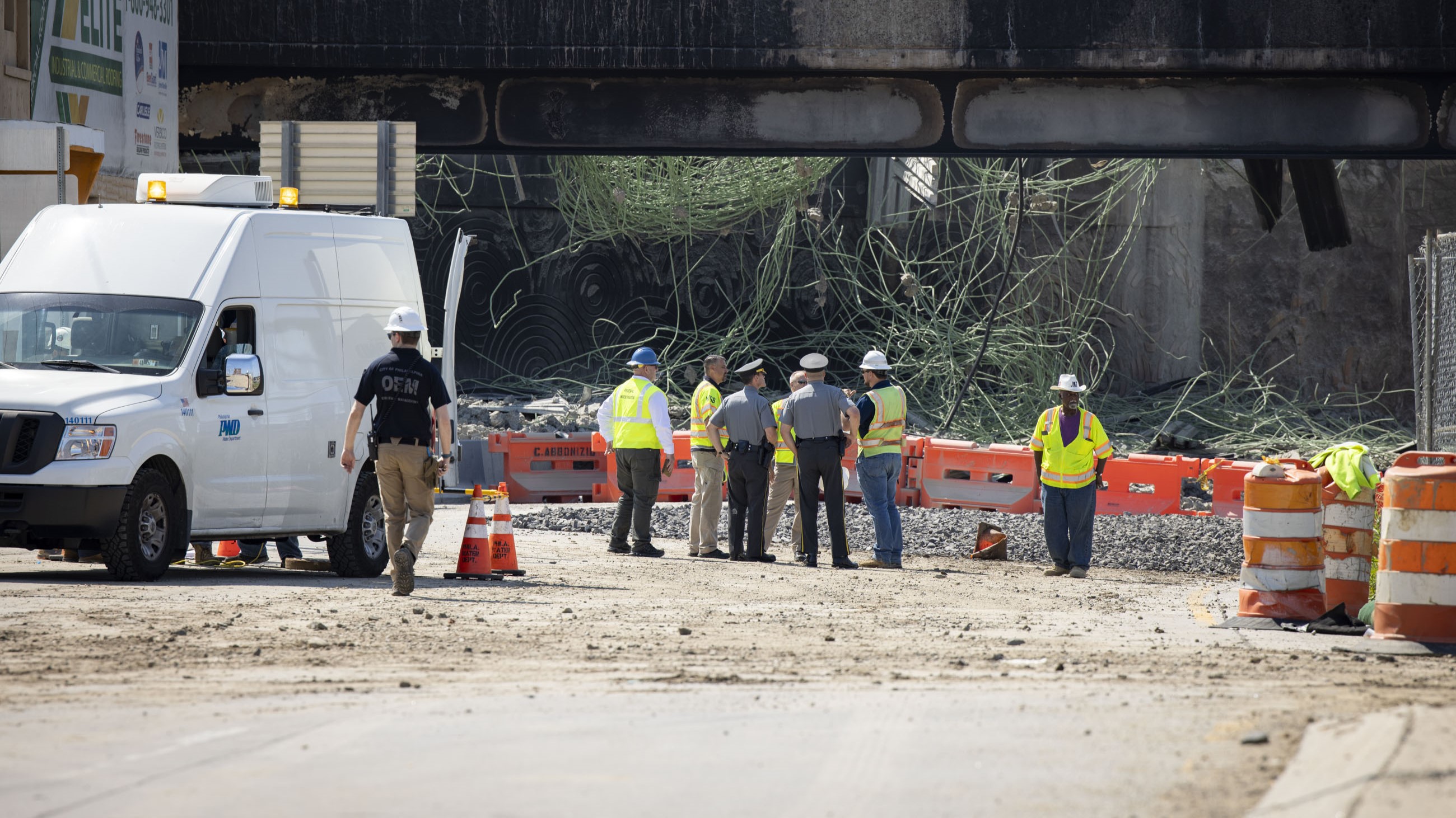 Pennsylvania State Police officers and several individuals wearing hard hats and yellow safety vests stand conversing near the site of the bridge collapse with orange barricades between them and the debris